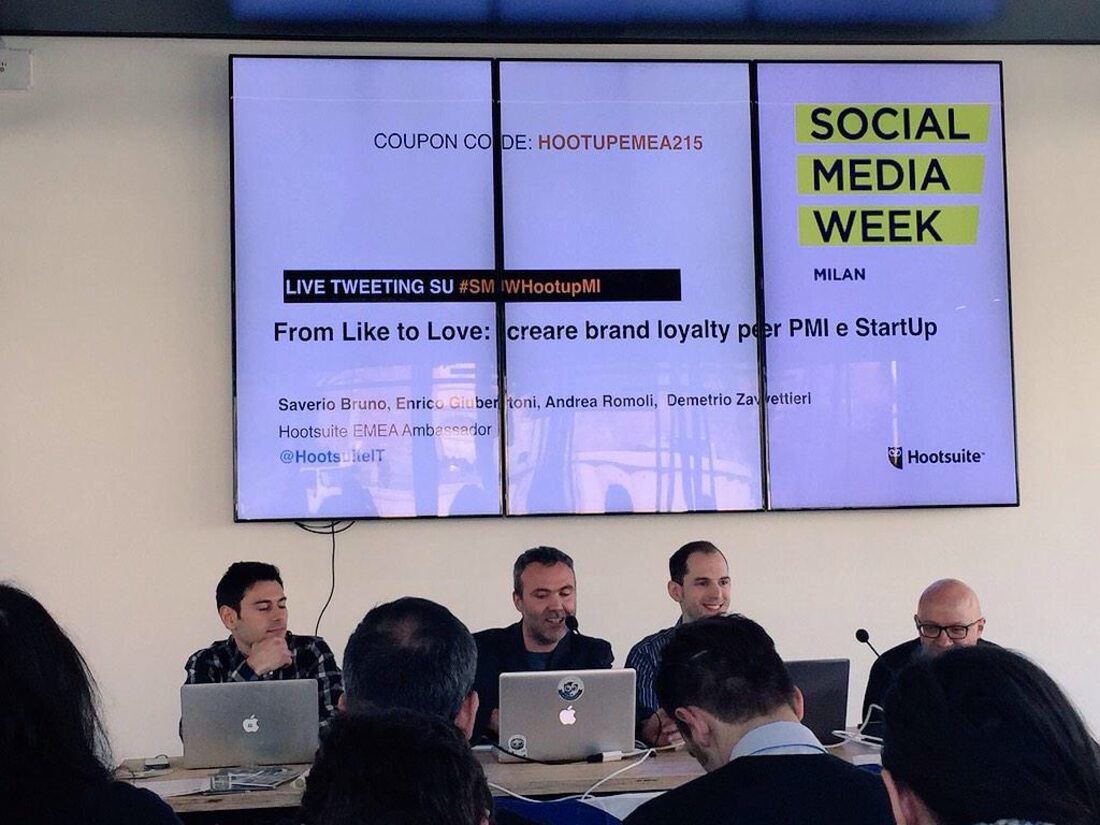 I quattro relatori al tavolo della Social Media Week Milano 2015: sullo schermo il titolo «From Like to Love: creare brand loyalty per PMI e StartUp» con i nomi di Saverio Bruno, Enrico Giubertoni, Andrea Romoli e Demetrio Zavettieri, ambasciatori Hootsuite per l’area europea